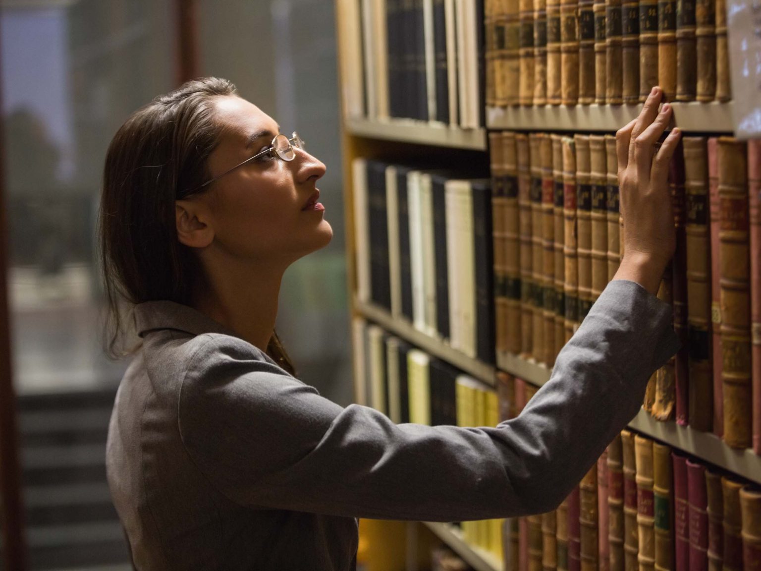 A woman standing in a library and looking through books on a shelf.